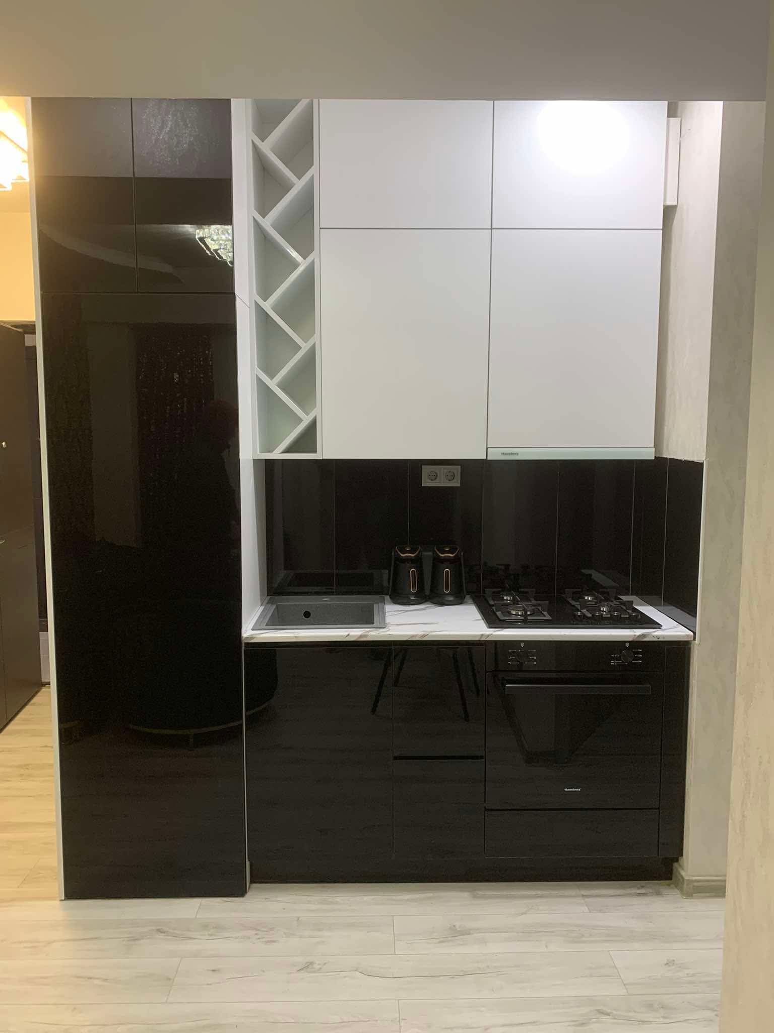 Compact modern kitchen with contrasting black high-gloss lower cabinets and appliances, white upper cabinets, and a white geometric wine rack. This small kitchen in Tbilisi features a built-in stovetop and oven, a stainless steel sink, and a black tiled backsplash.