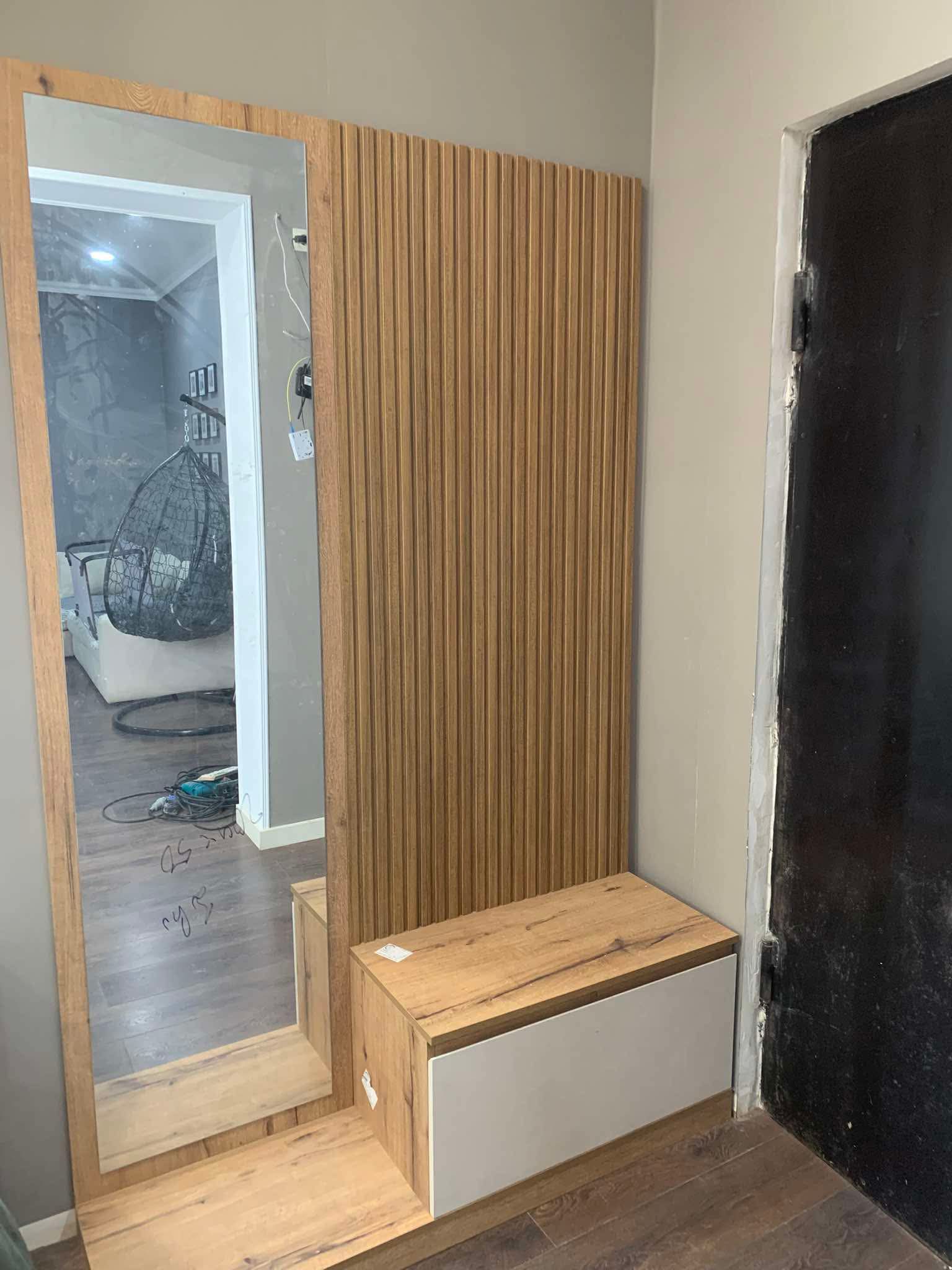 A modern entryway setup featuring a tall, rectangular mirror with a light wood frame leaning against a light gray wall. To the right of the mirror is a matching light wood panel with vertical fluting, and beneath it, a low wooden bench with a white drawer. The floor is dark wood, and the reflection in the mirror shows an interior space with a hanging egg chair.