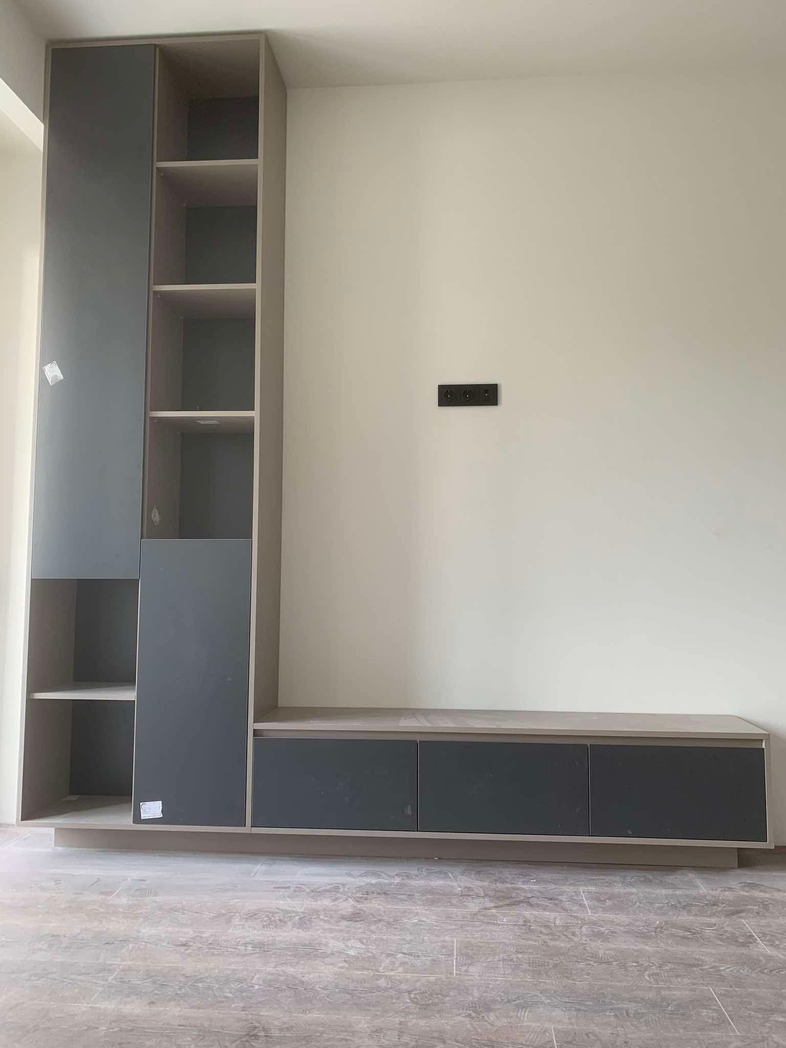 A modern, built-in entertainment unit or shelving system featuring a tall, narrow cabinet with both open shelves and closed gray doors on the left, connected to a long, low console with three gray drawers on the right. The unit is made of light wood with gray accents and stands against a plain cream-colored wall with an electrical outlet visible. The floor is light-colored and tiled.