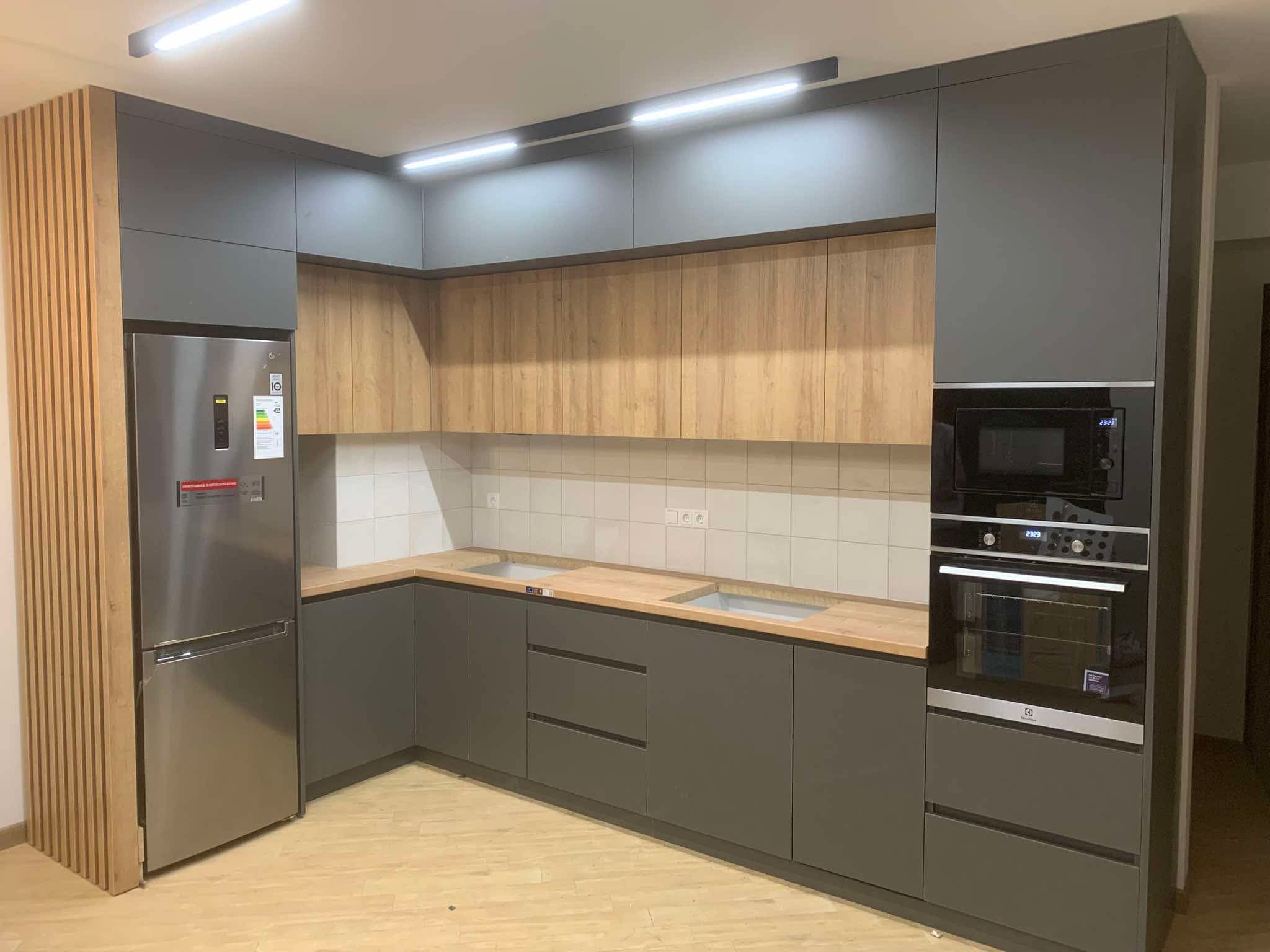Spacious modern kitchen featuring a combination of dark grey MDF (Medijapan) lower and upper cabinets, natural wood tone upper cabinets, and a matching wooden panel for the integrated refrigerator. This U-shaped kitchen includes built-in appliances, a light-tiled backsplash, and under-cabinet lighting, offering ample storage and counter space.