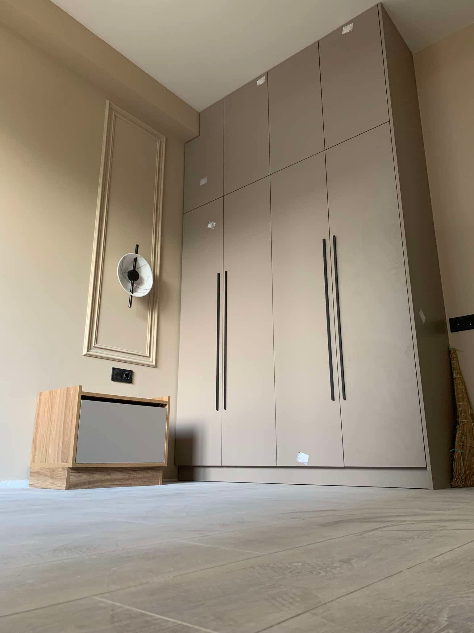 A modern bedroom featuring a large, floor-to-ceiling wardrobe with light brown doors and long black handles. To the left of the wardrobe, there is a wooden bedside table with a gray drawer. Above the bedside table, a decorative framed panel with a circular wall-mounted light fixture is visible on the light beige wall. The room has light wood flooring.